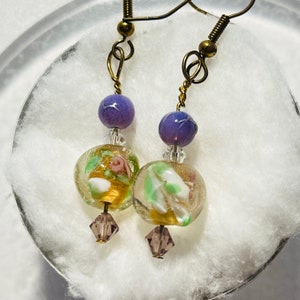 May include: A pair of dangle earrings with a gold tone wire and a purple glass bead, a clear glass bead with floral designs, and a crystal bead.