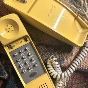 May include: A yellow GTE Automatic Electric telephone with a coiled cord. The handset features a gray keypad with numbers, letters, and symbols, and a label reading MAY 1978 8 5. The base unit has a matching yellow finish and a coiled cord.