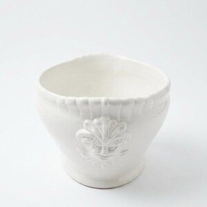 May include: A white ceramic bowl featuring a raised relief design of a face centered on the exterior. The upper rim of the bowl has a scalloped texture, and the overall finish is a glossy, solid white glaze with a wide, open circular opening.