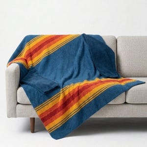 Puede incluir: Una manta azul con un diseño de rayas en tonos naranja, amarillo y rojo está sobre un sofá gris. La manta tiene una textura suave y un patrón clásico de inspiración suroeste. El sofá tiene patas de madera.