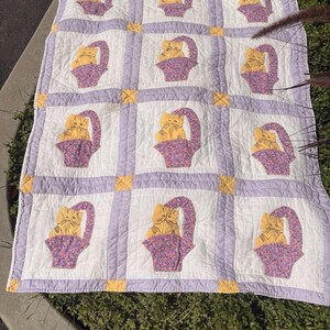 May include: A quilted blanket featuring a repeating pattern of yellow cats sitting in purple floral baskets. The quilt is constructed with white squares separated by light purple sashing and small yellow corner squares, creating a grid layout.