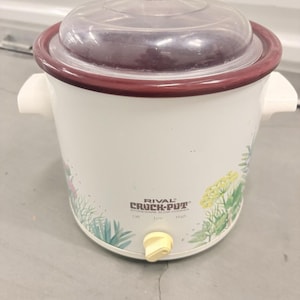 May include: A white Rival Crock-Pot stoneware slow cooker with a clear glass lid, two white side handles, and a yellow control knob. The exterior features a botanical print with green and yellow herbs. The text reads RIVAL CROCK-POT.