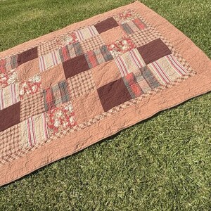 Puede incluir: Una colcha de retazos rectangular con un borde sólido de color rosa polvoriento y una cuadrícula central de cuadrados de tela alternos. Los cuadrados muestran varios patrones, incluyendo estampados florales, rayas verticales y texturas marrón oscuro.