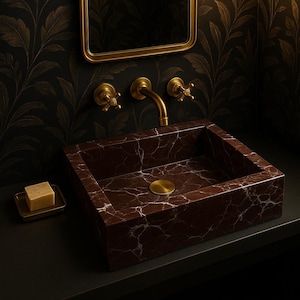 Op de afbeelding: Een donkerbruine marmeren wastafel met een gouden kraan en afvoerstop. De wastafel is gemonteerd op een zwart aanrecht in een badkamer met goudkleurig behang.