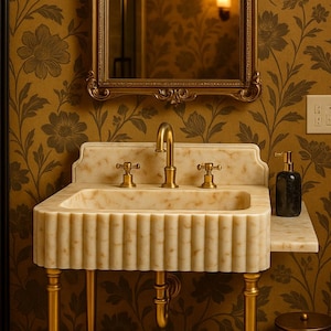 May include: A white marble bathroom sink with gold-toned faucets and legs. The sink is set against a wall with gold floral wallpaper and a gold-framed mirror.