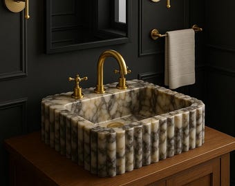 May include: A marble bathroom sink with a gold faucet and two knobs. The sink is on a wooden vanity with two cabinet doors. There are two gold wall sconces with white lampshades on either side of a black framed mirror.