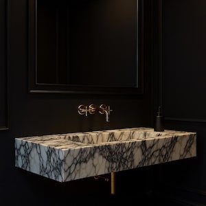 May include: A modern bathroom sink made of marble with gray veining, mounted on a black wall. The sink has a rectangular shape and is paired with gold-colored fixtures. A large black framed mirror hangs above the sink.