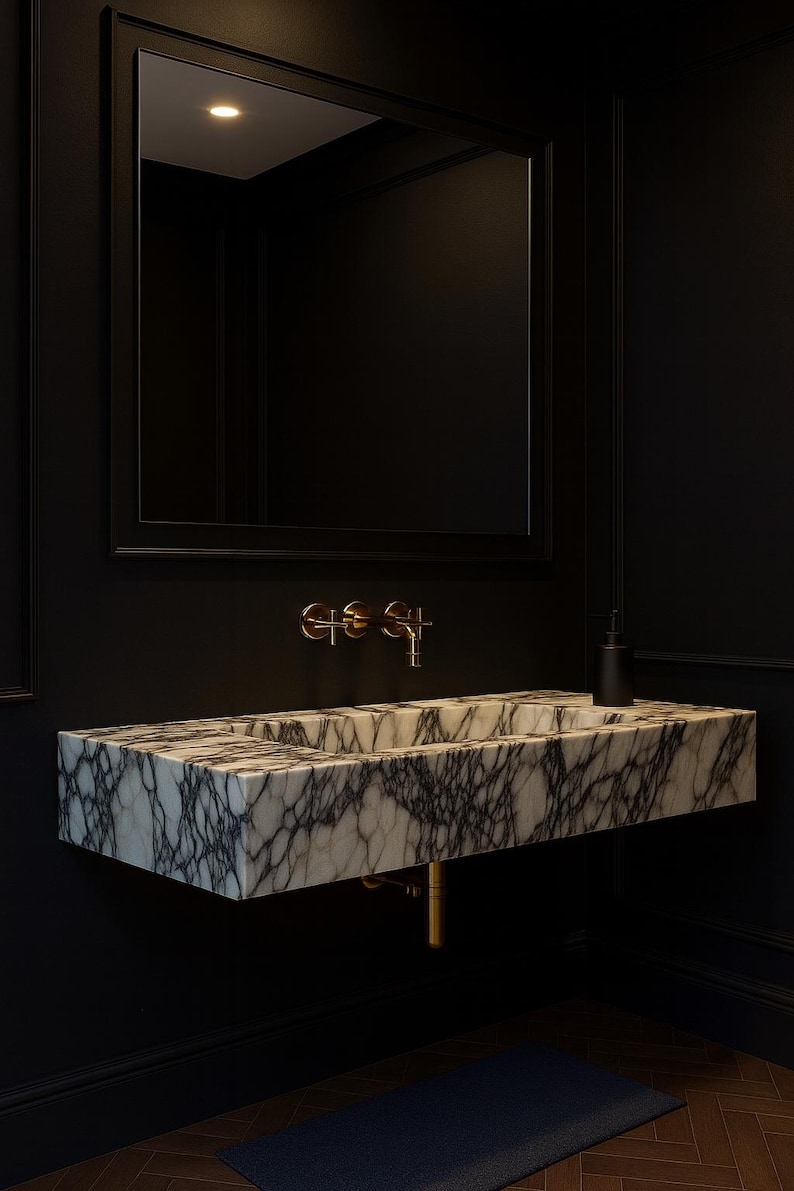 May include: A modern bathroom sink made of marble with gray veining, mounted on a black wall. The sink has a rectangular shape and is paired with gold-colored fixtures. A large black framed mirror hangs above the sink.