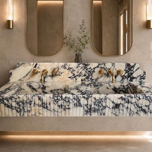 May include: A marble double sink with gold taps. The sink has a white and black veined pattern and is mounted on a neutral-toned base. Two oval mirrors and sconces are mounted on the wall above the sink.