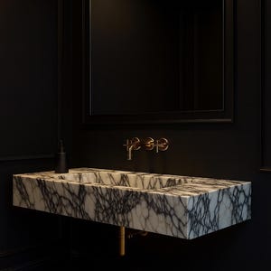 May include: A modern bathroom sink made of marble with gray veining, mounted on a black wall. The sink has a rectangular shape with a built-in basin. Gold-colored faucet fixtures and a black soap dispenser are visible. A black framed mirror hangs above the sink.