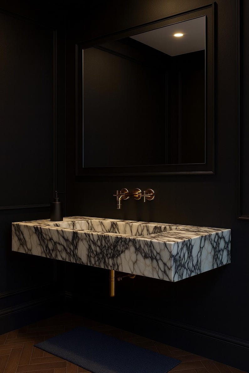 May include: A modern bathroom sink made of marble with gray veining, mounted on a black wall. The sink has a rectangular shape with a built-in basin. Gold-colored faucet fixtures and a black soap dispenser are visible. A black framed mirror hangs above the sink.