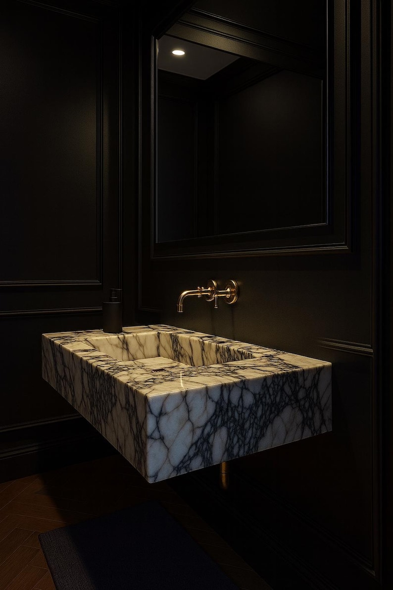 May include: A modern bathroom sink made of marble with gray veining. The sink is rectangular and mounted on a dark wall. A gold faucet and a black soap dispenser are mounted above the sink. A mirror is above the sink.