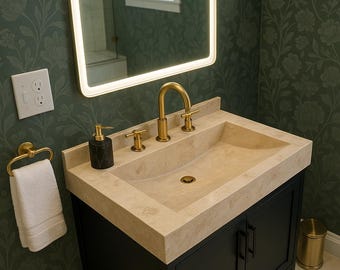 May include: A black bathroom vanity with a white countertop and a gold faucet. The vanity has two doors and a single sink. The sink is made of a light-colored stone. The vanity is in a bathroom with green floral wallpaper.