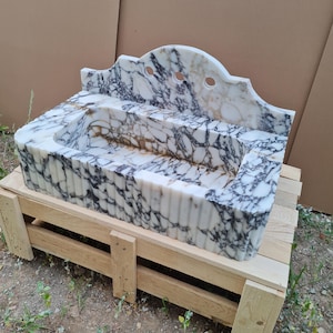 May include: A white marble sink with black veining, featuring a scalloped backsplash with three holes. The sink is rectangular with a tiered design and sits on a wooden pallet. The marble has a polished finish.