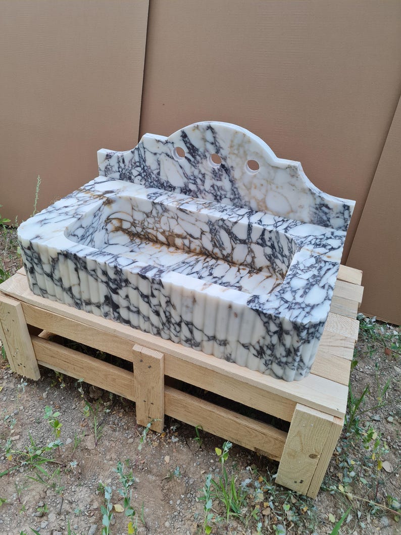 May include: A white marble sink with black veining, featuring a scalloped backsplash with three holes. The sink is rectangular with a tiered design and sits on a wooden pallet. The marble has a polished finish.