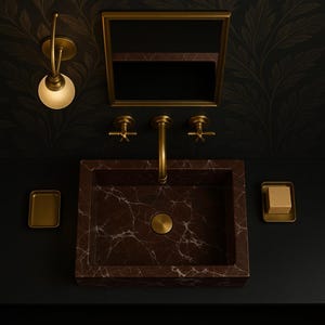 Op de afbeelding: Een bruine marmeren wastafel met een gouden kraan en twee gouden handvatten. De wastafel is geplaatst tegen een zwarte achtergrond met een gouden spiegel erboven.