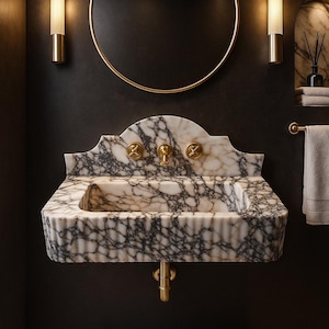 May include: A luxurious bathroom sink made of marble with black veining. The sink has gold fixtures and is mounted on a dark wall. A round gold mirror and sconces flank the sink, creating a sophisticated and elegant aesthetic.