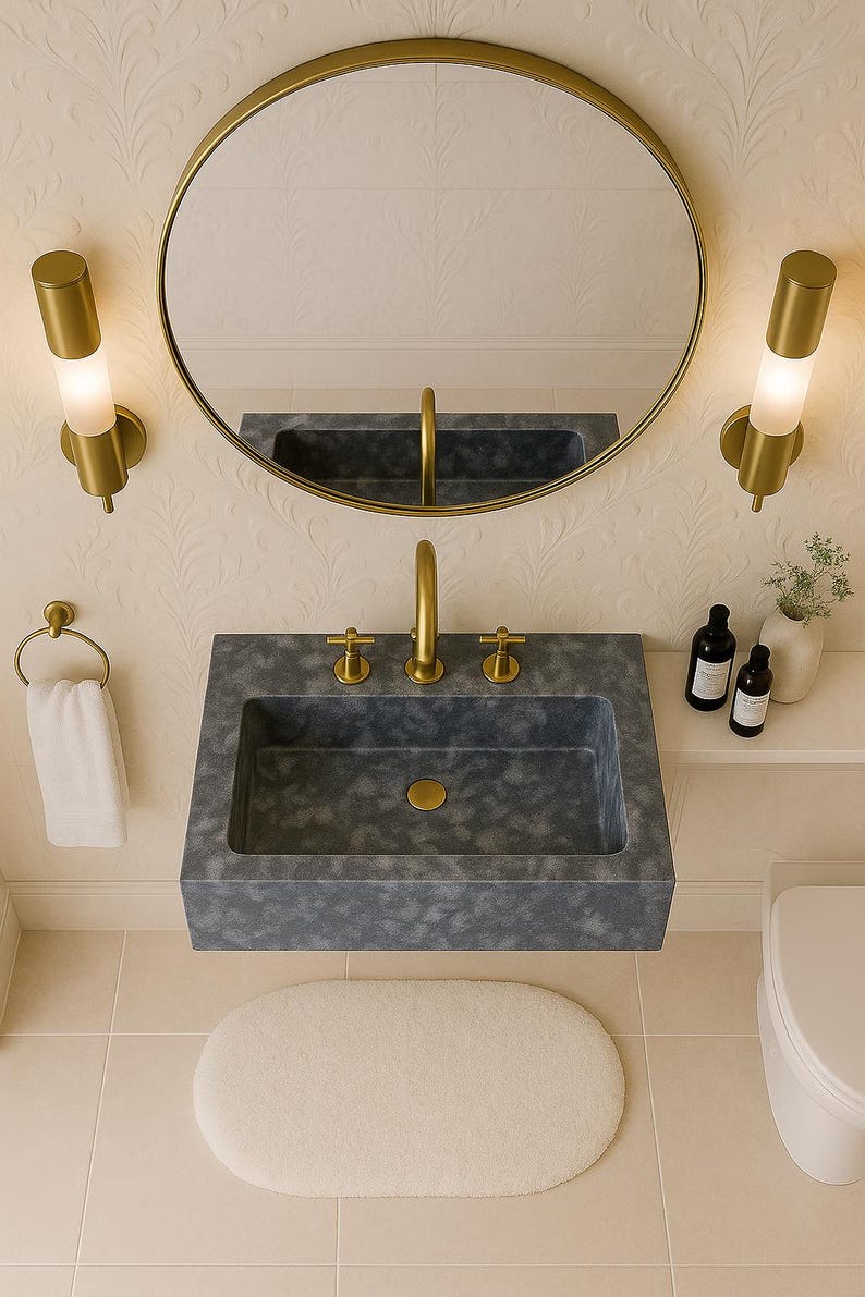 May include: A contemporary bathroom suite featuring a grey rectangular sink with gold taps and a circular gold mirror. Two gold wall lights are either side of the mirror. An oval white rug is on the floor. The walls have a floral pattern.