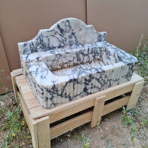 May include: A marble sink with a white and black veined pattern. The sink has a decorative, arched backsplash with three circular cutouts. It rests on a wooden pallet, showcasing its size and craftsmanship. The sink's basin is rectangular with a fluted front.