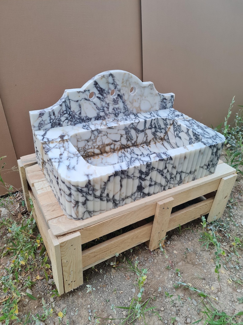 May include: A marble sink with a white and black veined pattern. The sink has a decorative, arched backsplash with three circular cutouts. It rests on a wooden pallet, showcasing its size and craftsmanship. The sink's basin is rectangular with a fluted front.
