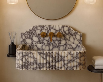 May include: A marble sink with a gray and white pattern, gold fixtures, and a matching mirror. Two pendant lights with ribbed glass hang on either side of the mirror. A dark brown rug sits on the floor.