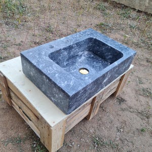 May include: A rectangular, grey marble sink with a central drain hole. The sink rests on a wooden crate, highlighting its natural stone texture and design. Sink dimensions are not visible.
