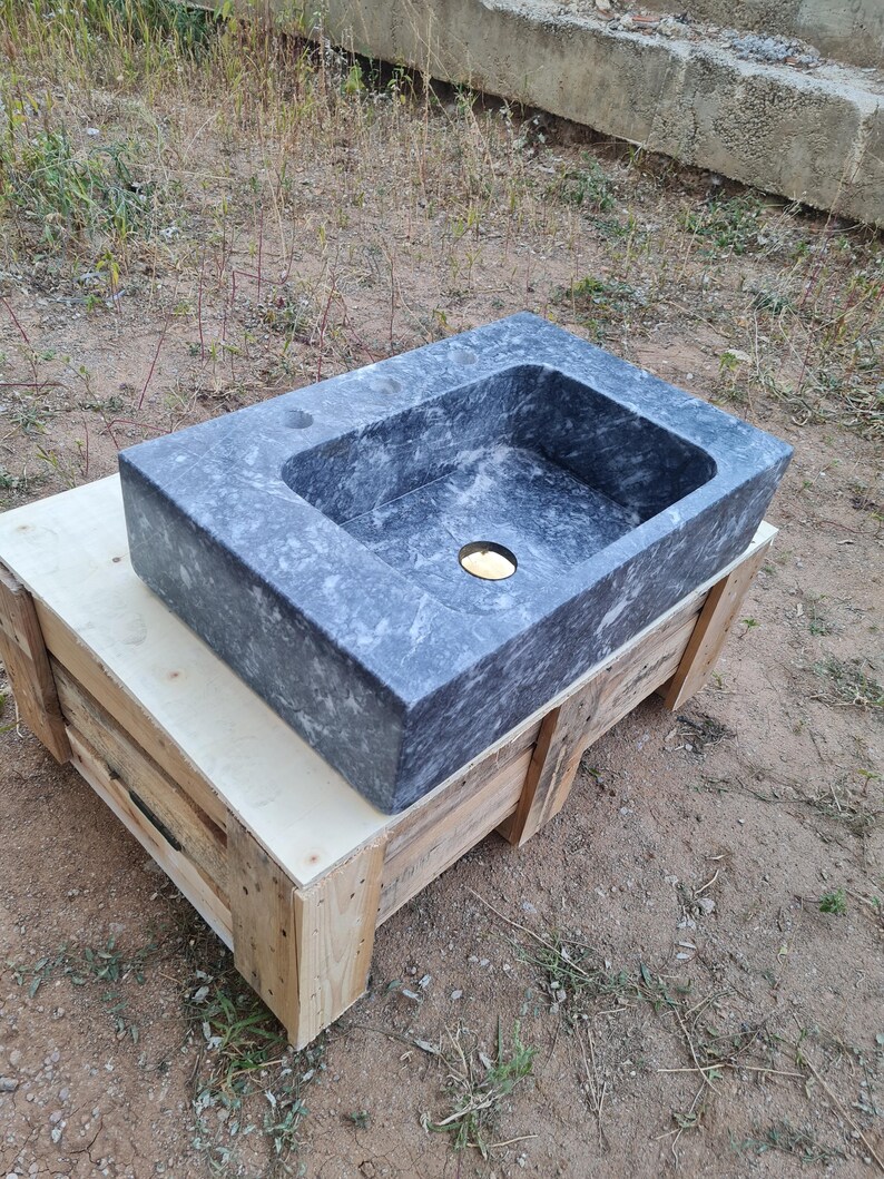 May include: A rectangular, grey marble sink with a central drain hole. The sink rests on a wooden crate, highlighting its natural stone texture and design. Sink dimensions are not visible.