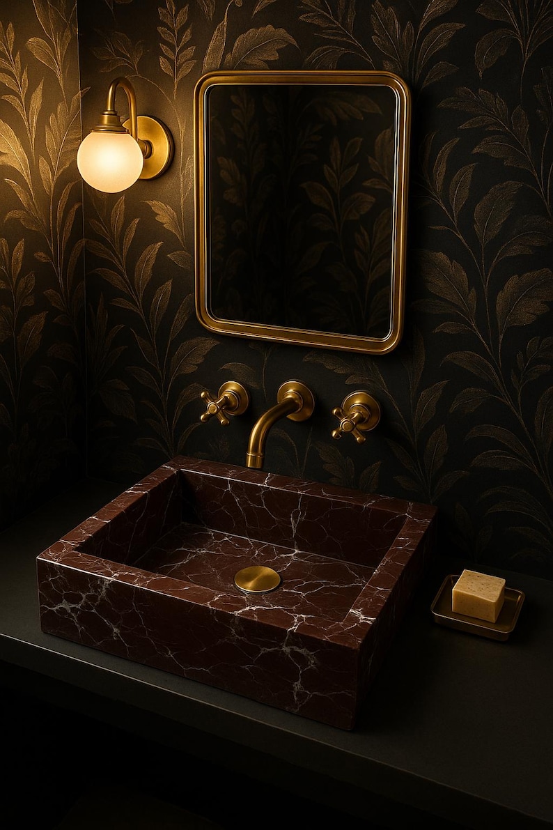 Op de afbeelding: Een donkerbruine marmeren wastafel met een gouden kraan en twee gouden knoppen. De wastafel is geplaatst tegen een zwarte muur met goudkleurig behang.
