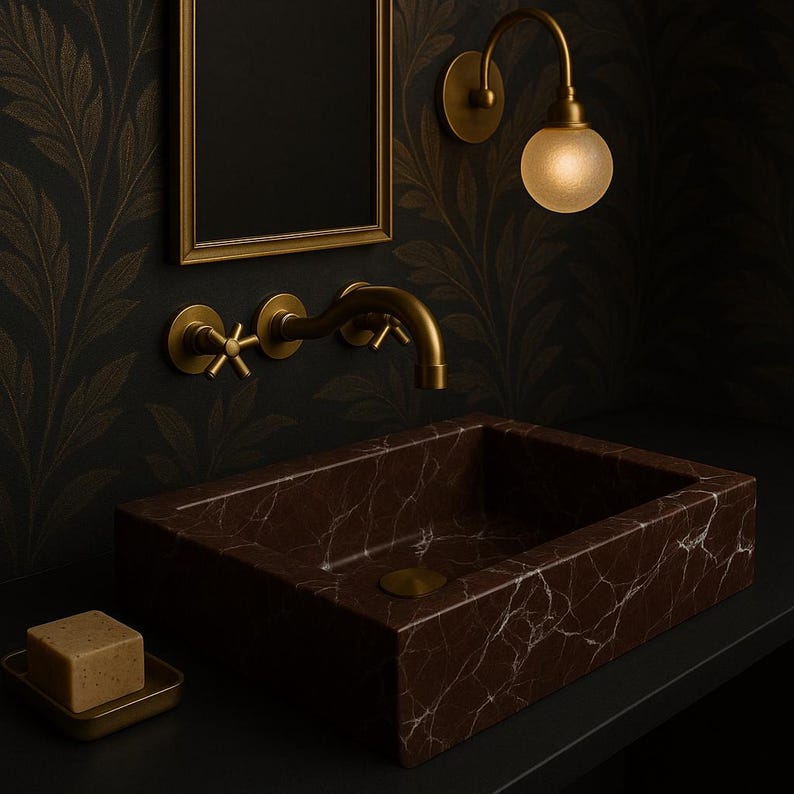 Op de afbeelding: Een bruine marmeren badkamer wastafel met een messing kraan en een messing wandlamp met een matglazen bol. De wastafel staat op een zwart aanrecht.