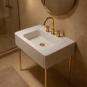 May include: A white marble bathroom sink with a fluted design and gold-colored legs. The sink has a gold faucet and fixtures. A round gold mirror and a wall-mounted light fixture are visible in the background.