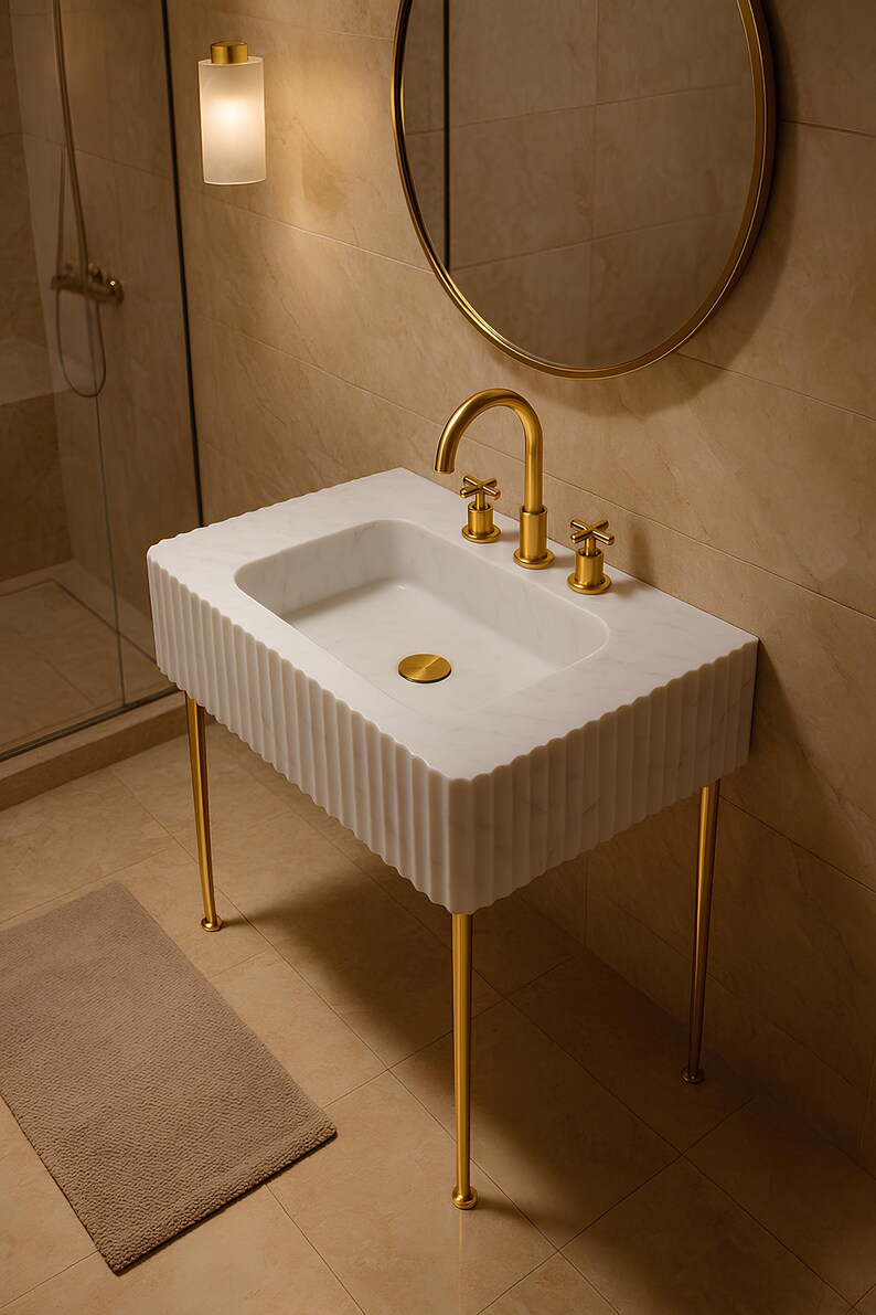 May include: A white marble bathroom sink with a fluted design and gold-colored legs. The sink has a gold faucet and fixtures. A round gold mirror and a wall-mounted light fixture are visible in the background.