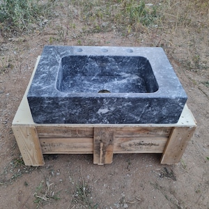 May include: A rectangular, dark grey marble sink with three tap holes and a central drain. The sink is set on a wooden pallet. The marble has a natural, veined pattern. The sink is outdoors.