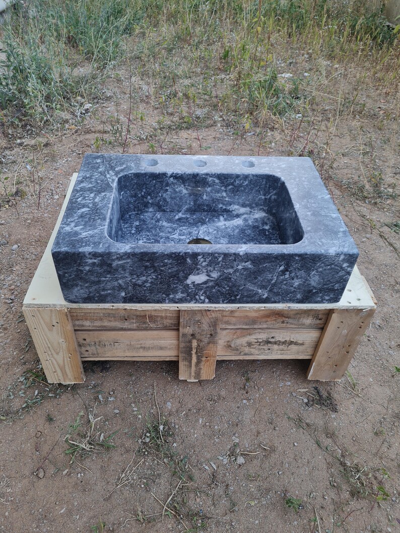 May include: A rectangular, dark grey marble sink with three tap holes and a central drain. The sink is set on a wooden pallet. The marble has a natural, veined pattern. The sink is outdoors.