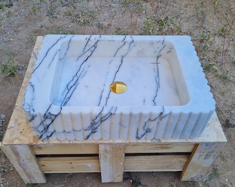 May include: A rectangular white marble sink with black veining and a gold-colored drain. The sink has a fluted edge and is set on a wooden pallet. The sink is a bathroom fixture.