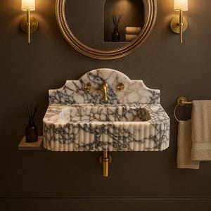 May include: A luxurious bathroom sink made of white marble with gray veining. The sink has a fluted design and gold fixtures, including a faucet and drainpipe. A round gold mirror hangs above the sink, and two sconces flank it. A beige bath mat is on the floor.