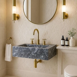 May include: A contemporary bathroom with a grey marble sink and gold taps, a circular mirror, and wall lights. A white towel hangs on a gold ring. The walls have a floral design, and a white oval rug is on the floor.