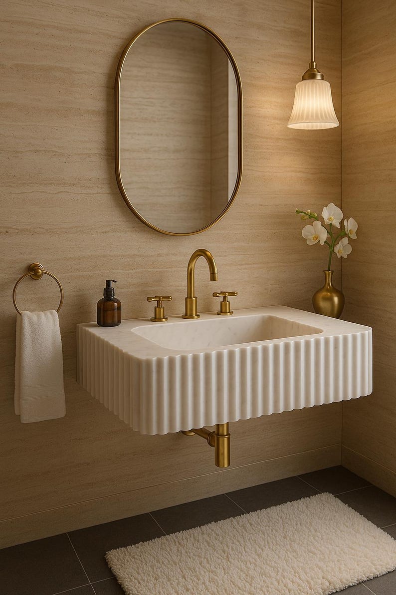 May include: A modern bathroom sink with a fluted white marble basin and gold fixtures. The oval mirror and pendant light complement the design. A white towel hangs on a gold ring, and a small vase holds white flowers.