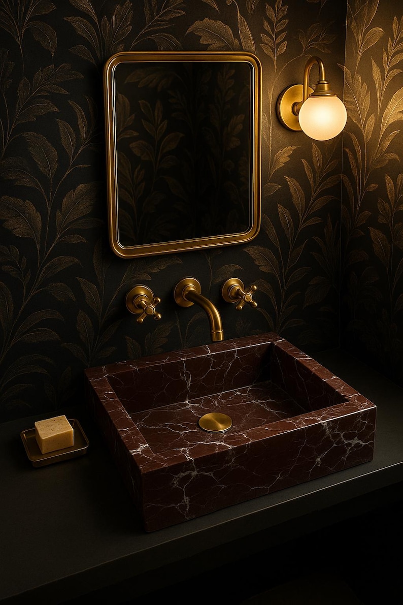 Op de afbeelding: Een donkerbruine marmeren wastafel met een gouden kraan en afvoerstop. De wastafel is gemonteerd op een zwart aanrecht in een badkamer met goudkleurig behang.