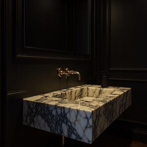 May include: A modern bathroom sink made of marble with dark gray veining. The sink is mounted on a black wall, with a gold faucet and a black soap dispenser. A mirror is above the sink.