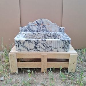 May include: A marble sink with a white and black veined pattern. The sink has a decorative back with three circular cutouts. It is set on a wooden pallet. The sink's basin has a ribbed design.