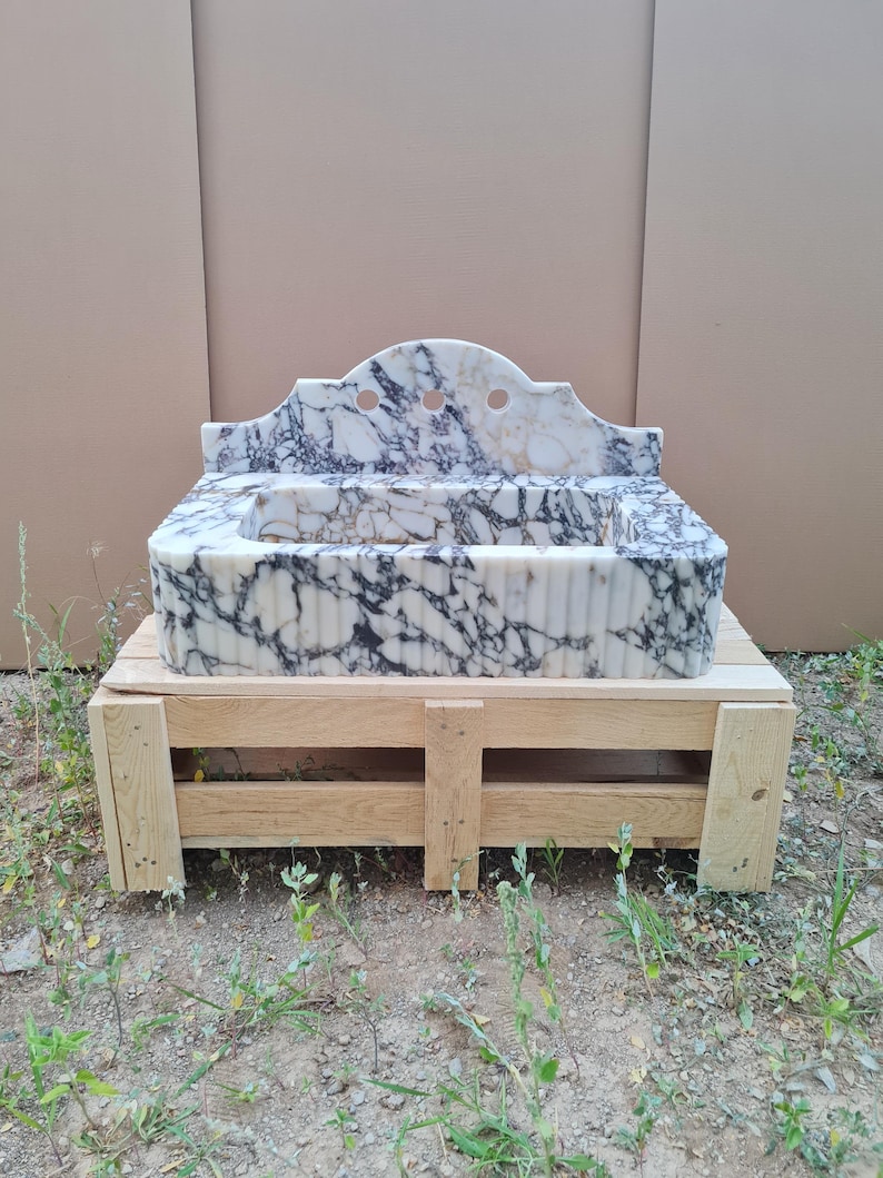 May include: A marble sink with a white and black veined pattern. The sink has a decorative back with three circular cutouts. It is set on a wooden pallet. The sink's basin has a ribbed design.