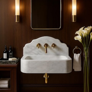 May include: An elegant bathroom interior with a white marble sink and gold taps. The room features a rectangular mirror, wall sconces, and a vase of white calla lilies. The dark wood paneling and a small side table with toiletries create a sophisticated atmosphere.