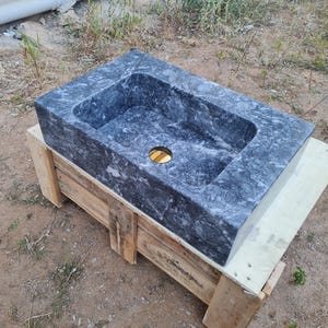 May include: A dark grey marble sink with a rectangular basin and a gold drain. The sink is displayed on a wooden pallet. The marble has a natural pattern. Suitable for bathroom renovations, the sink is made of stone.