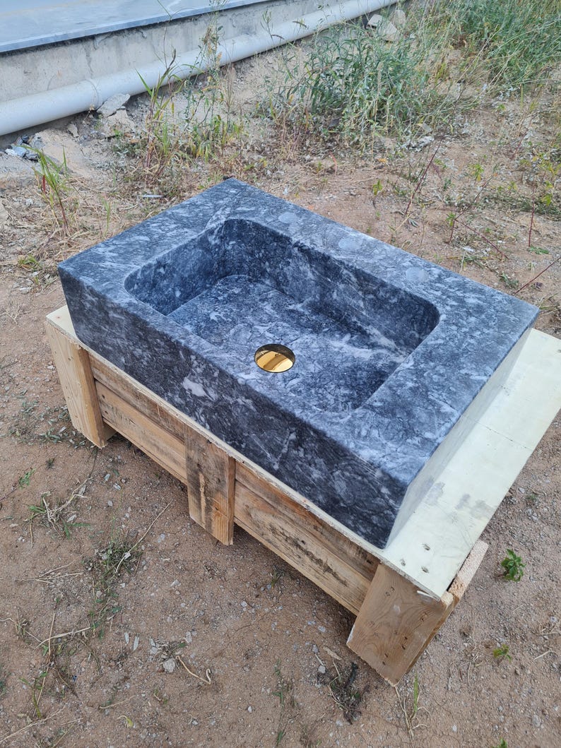 May include: A dark grey marble sink with a rectangular basin and a gold drain. The sink is displayed on a wooden pallet. The marble has a natural pattern. Suitable for bathroom renovations, the sink is made of stone.