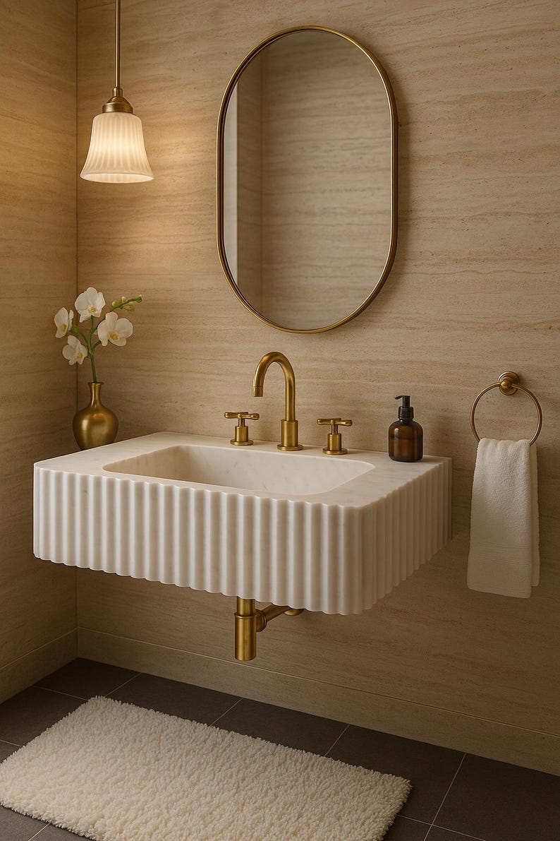 May include: A modern bathroom scene featuring a white marble sink with a fluted design, gold fixtures, and an oval mirror. A small vase with white flowers and a pendant light complete the elegant aesthetic.
