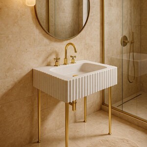 May include: A white, fluted bathroom sink with gold-colored fixtures and legs. A round mirror and shower enclosure are visible in the background. The sink has a rectangular basin and a gold faucet.