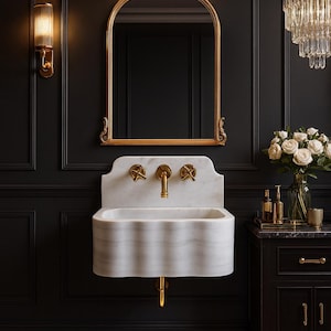 May include: A luxurious bathroom scene featuring a white marble sink with gold fixtures, set against a dark wall. A gold-framed mirror hangs above the sink, with a chandelier and sconces adding to the elegant ambiance. A fluffy white rug sits on the floor.