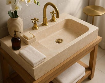 May include: A beige rectangular bathroom sink with a gold faucet and fixtures, set on a wooden vanity. A brown soap dispenser, soap dish, and folded white towel are on the counter. A vase of white orchids adds a decorative touch.
