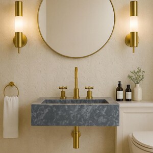 May include: A contemporary bathroom design with a grey stone sink and gold taps, a circular gold mirror, and matching wall lights. A white towel hangs on a gold ring. The walls have a subtle floral pattern, and a small rug sits on the floor.