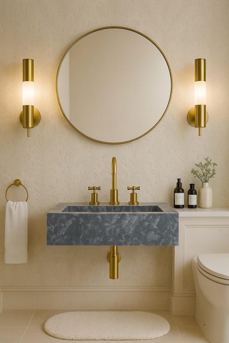 May include: A contemporary bathroom design with a grey stone sink and gold taps, a circular gold mirror, and matching wall lights. A white towel hangs on a gold ring. The walls have a subtle floral pattern, and a small rug sits on the floor.
